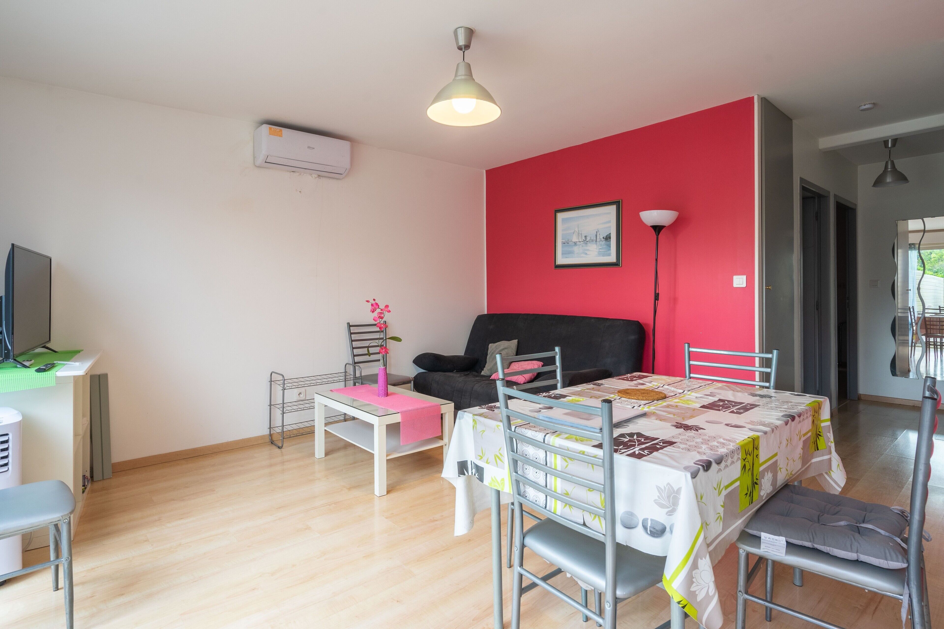 Ett rum med en röd vägg, en svart soffa, ett matbord med en duk, en metallstol, ett litet bord med en rosa duk, en golvlampa och en tv som står på ett stativ.