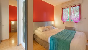 Un dormitorio con cama, mesita de noche, ventana con cortinas y puerta que da a otra habitación.
