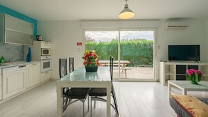 Eine moderne Küche mit weißen Schränken, einem Esstisch mit Stühlen, einem Fernseher und Blick auf Grün hinter einer Schiebetür.