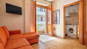 Un soggiorno con divano, televisore, lavatrice e una porta scorrevole che dà su un balcone.