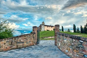 Uma trilha de pedra leva a um edifício histórico com uma torre de sino, cercado por vegetação e um céu nublado.