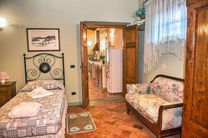 Quarto com cama, sofá e área de cozinha visível por uma porta aberta, além de um quadro na parede.