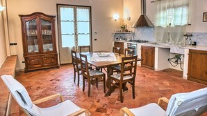 A dining area with a wooden table and chairs, a cabinet with glass doors, a window with curtains, and a kitchen area with a stove and sink. - Apartment 'Holiday Home In Tuscany' with Mountain View, Private Terrace and Wi-Fi (Castelfiorentino)