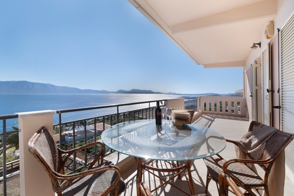 A balcony with a glass table and wicker chairs, overlooking a coastal view.