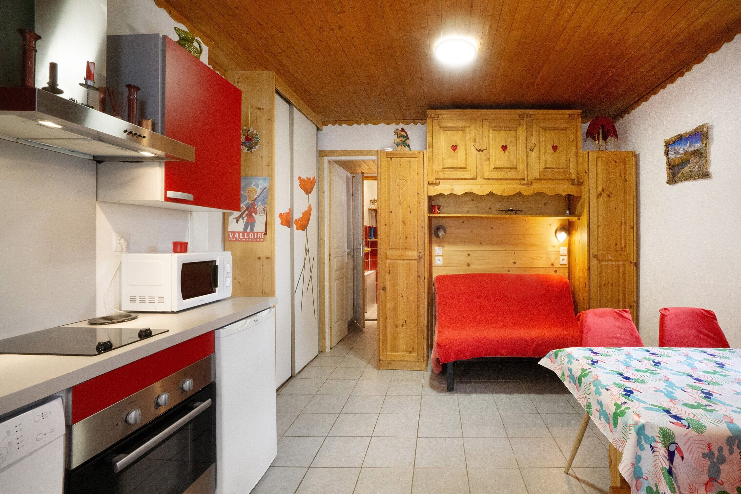 A compact kitchen with a microwave, stove, and oven, a wooden cabinet with a red bedspread, and a small table with a colorful tablecloth.