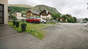A parking area with a red car and a white car parked in front of a building with a mountain backdrop. - Apartment 'Studio Cabine - Les Ecrins 2' with Mountain View and Wi-Fi (Valloire)