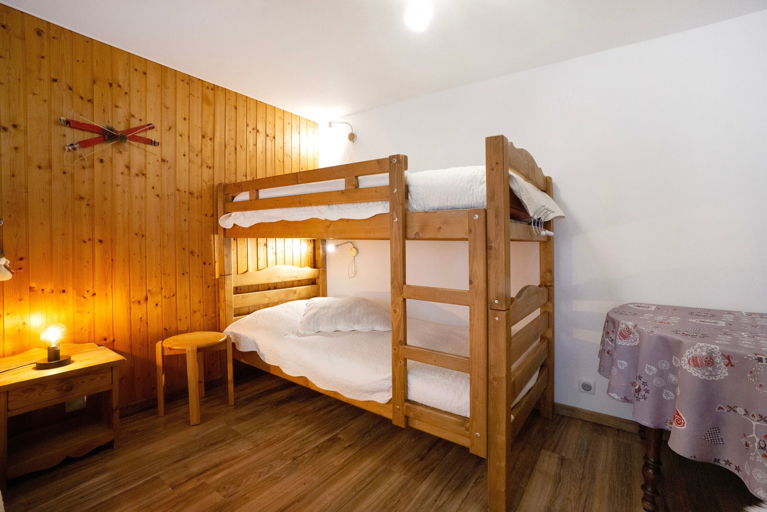 A wooden bunk bed room with a small table and chair, a wall-mounted ski rack, and a table with a patterned tablecloth.
