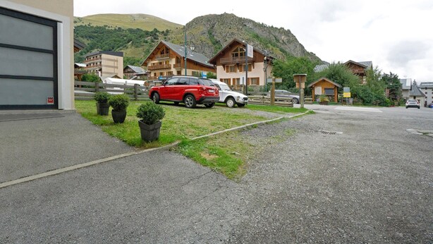 Uma área de estacionamento com um carro vermelho e um carro branco estacionados em frente a um edifício, com uma montanha ao fundo.
