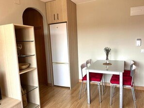 Kompakti keittiö, jossa on sisäänrakennettu jääkaappi, punaisilla tuoleilla varustettu ruokapöytä ja puinen hyllykkö.