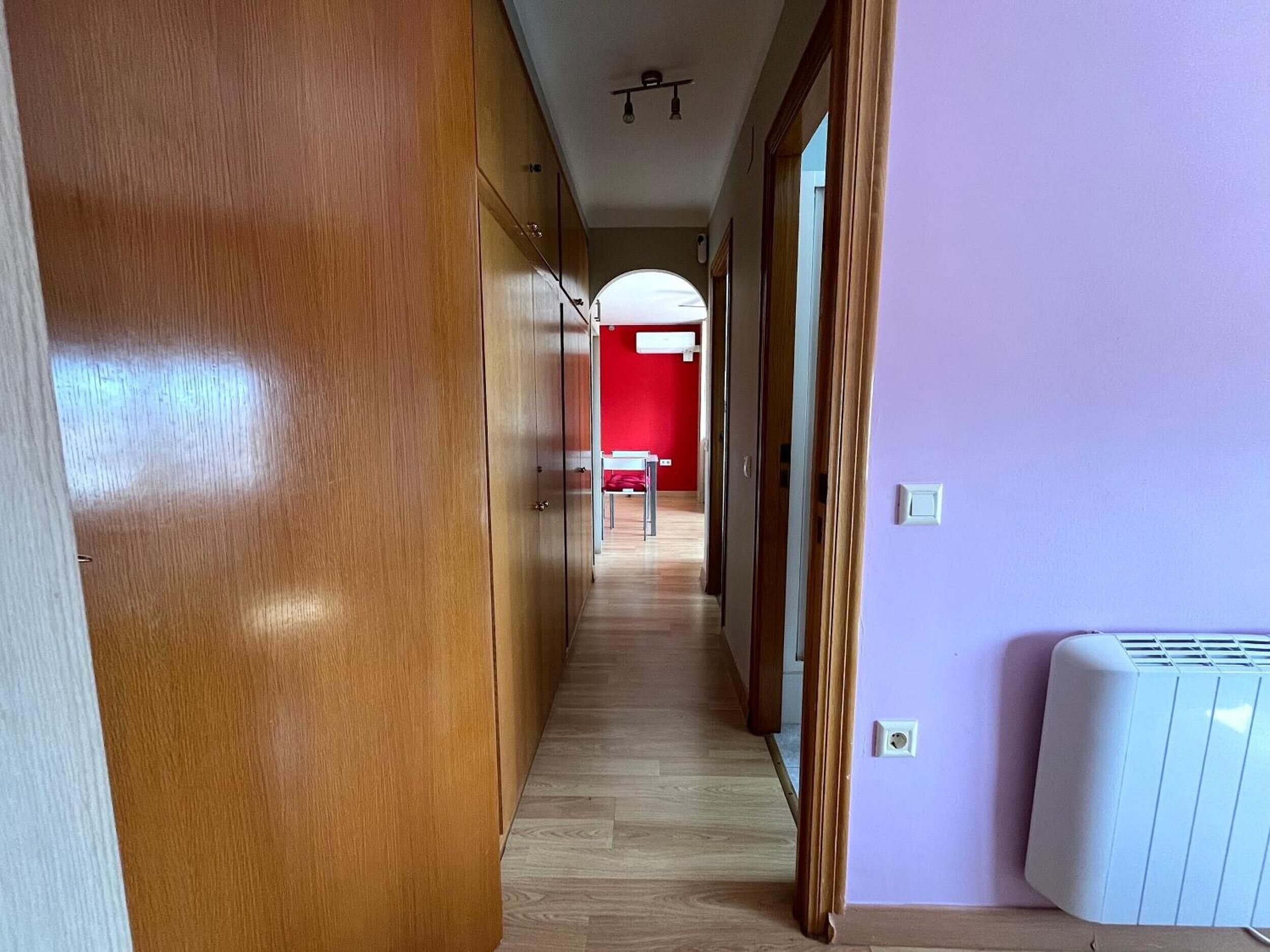 Un couloir avec un plancher en bois, un mur rouge au fond et un radiateur blanc sur la droite.