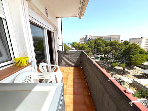 A balcony with a view of buildings and trees, a chair, a table, and a yellow basket.