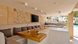 A modern outdoor dining area with a stone wall, a flat-screen TV, and a pool in the background. - Villa 'Oasis Del Mar - Splendid' with Sea View, Wi-Fi and Air Conditioning (IB)