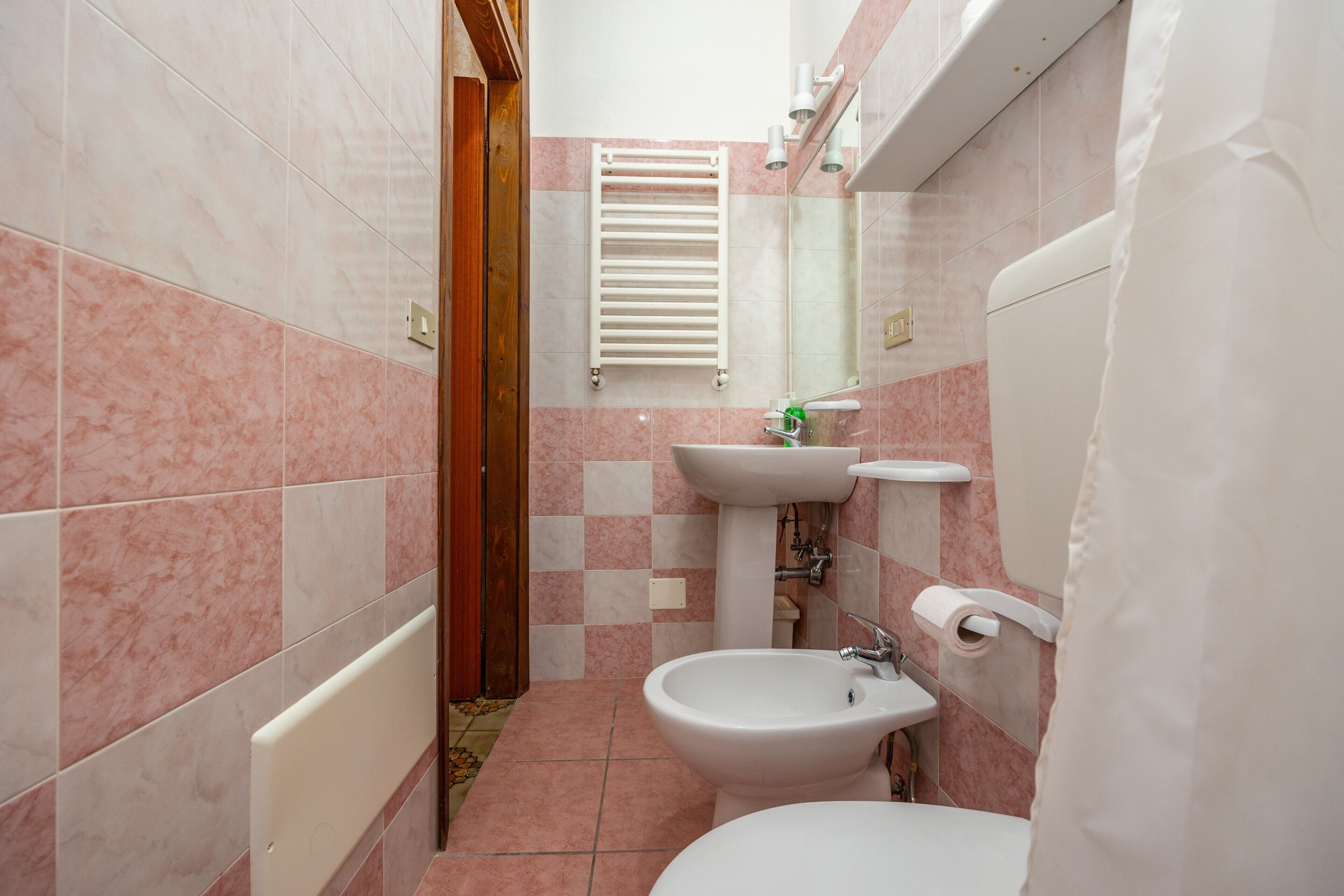 A bathroom with pink and white tiles, a white radiator, a bidet, a sink, and a toilet.