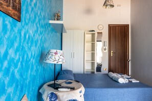 Une chambre avec un mur bleu à texture, un lit recouvert d’une couverture bleue, une lampe de chevet, une télécommande, une porte en bois et une armoire blanche.