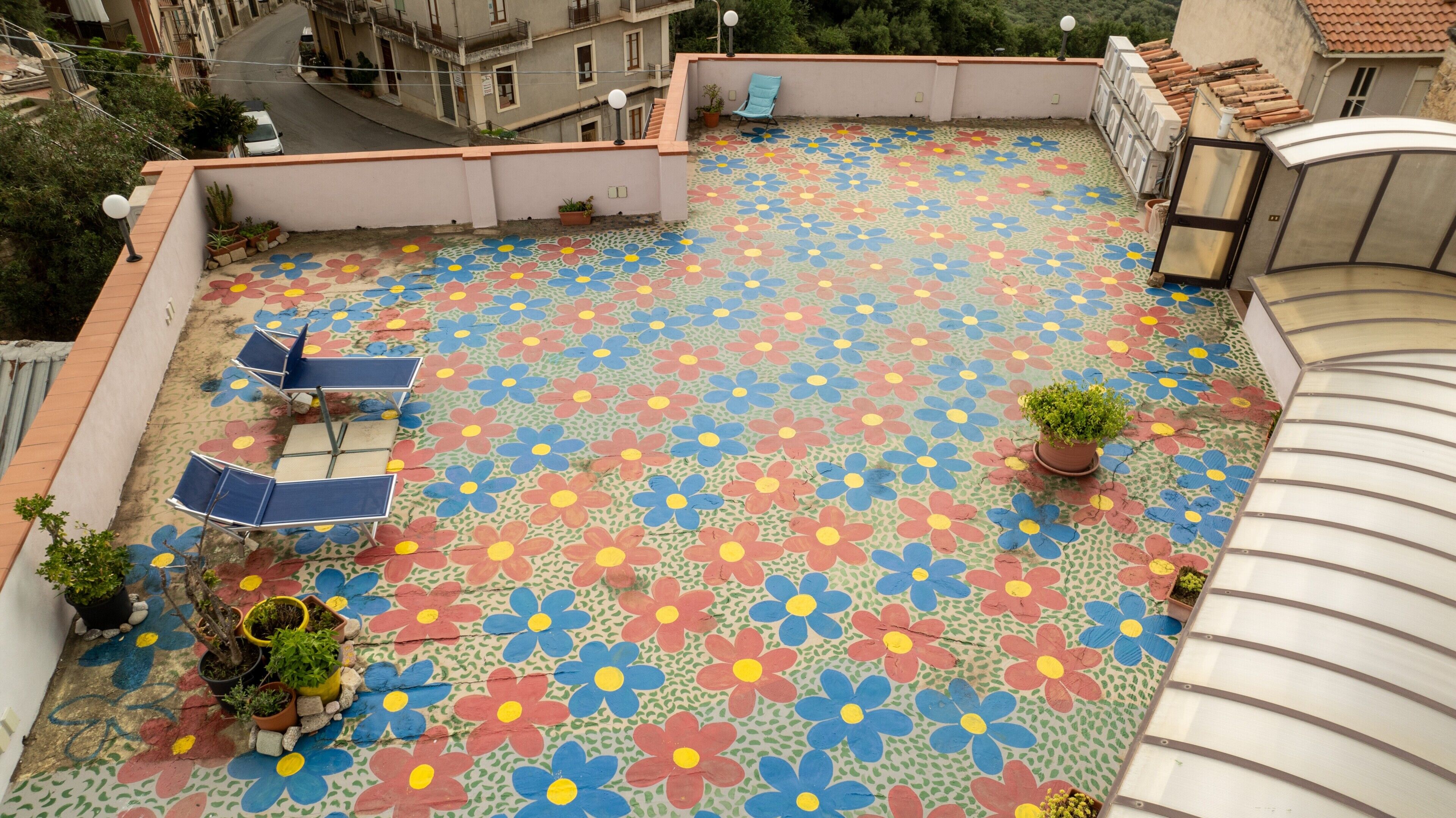 A rooftop terrace with a colorful flower-patterned floor, potted plants, and blue chairs.