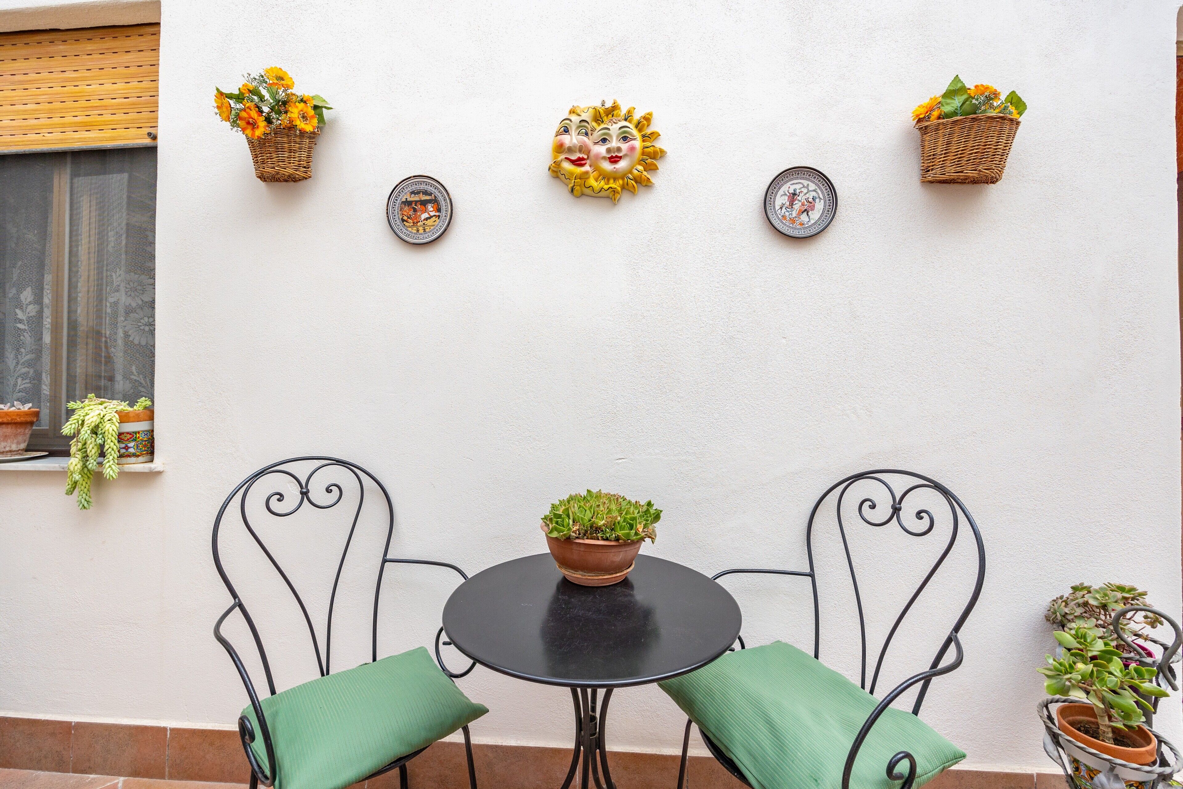 Un petit espace extérieur aménagé avec une table ronde et deux chaises, une plante en pot posée sur la table, et des paniers fixés au mur, contenant des fleurs et des assiettes décoratives.