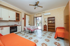 A compact room with a kitchenette, a dining table, a ceiling fan, and a bed with an orange bedspread.