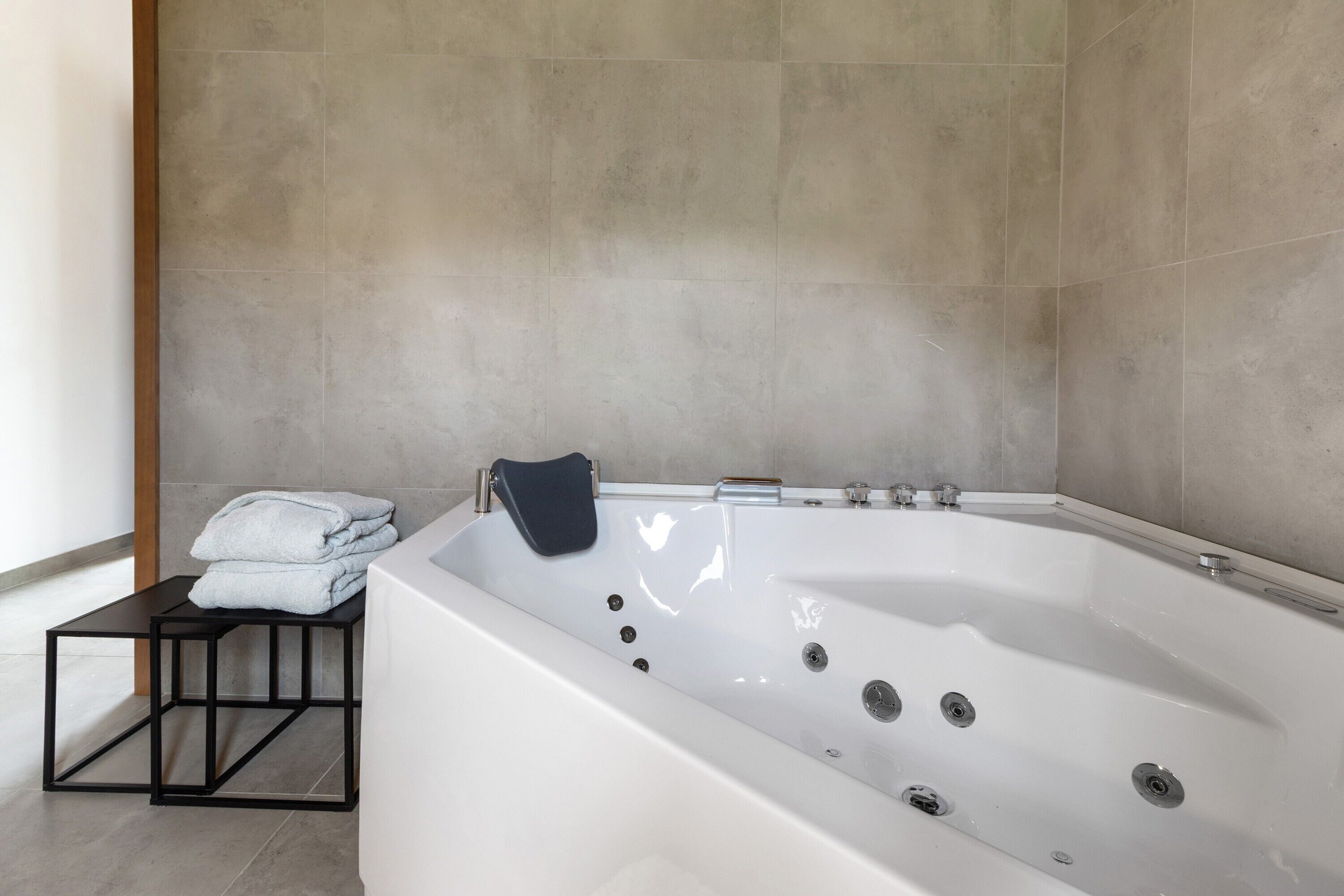 A modern bathroom with a large bathtub, a small black table with towels, and a plain tiled wall.