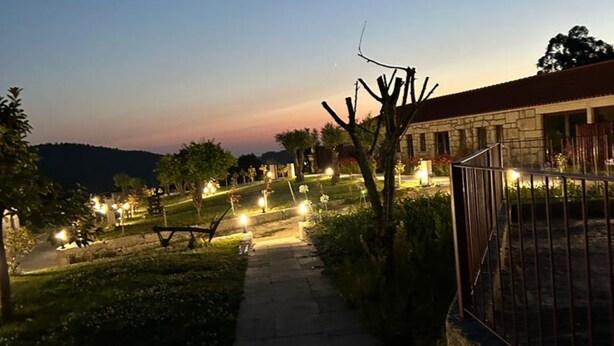 Un sendero conduce a un edificio de fachada de piedra, rodeado de jardines iluminados y un paisaje montañoso.