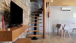 Un interior moderno con un mueble de televisión de madera, una pared de piedra, una escalera de madera y una zona de comedor con mesa y sillas.