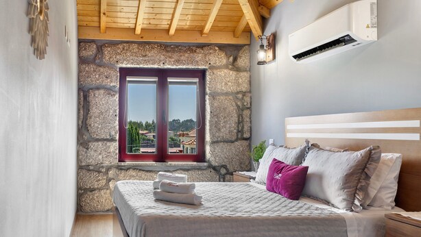 Un dormitorio con pared de piedra, una cama con almohadas, una ventana y un equipo de aire acondicionado.