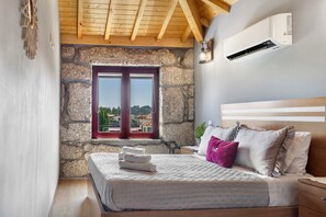 A bedroom with a stone wall, a bed with pillows, a window, and an air conditioning unit. - Quinta of Carvoeiras  'Casa Do Caseiro - T2' with Lake View, Wi-Fi and Air Conditioning (Vieira do Minho)