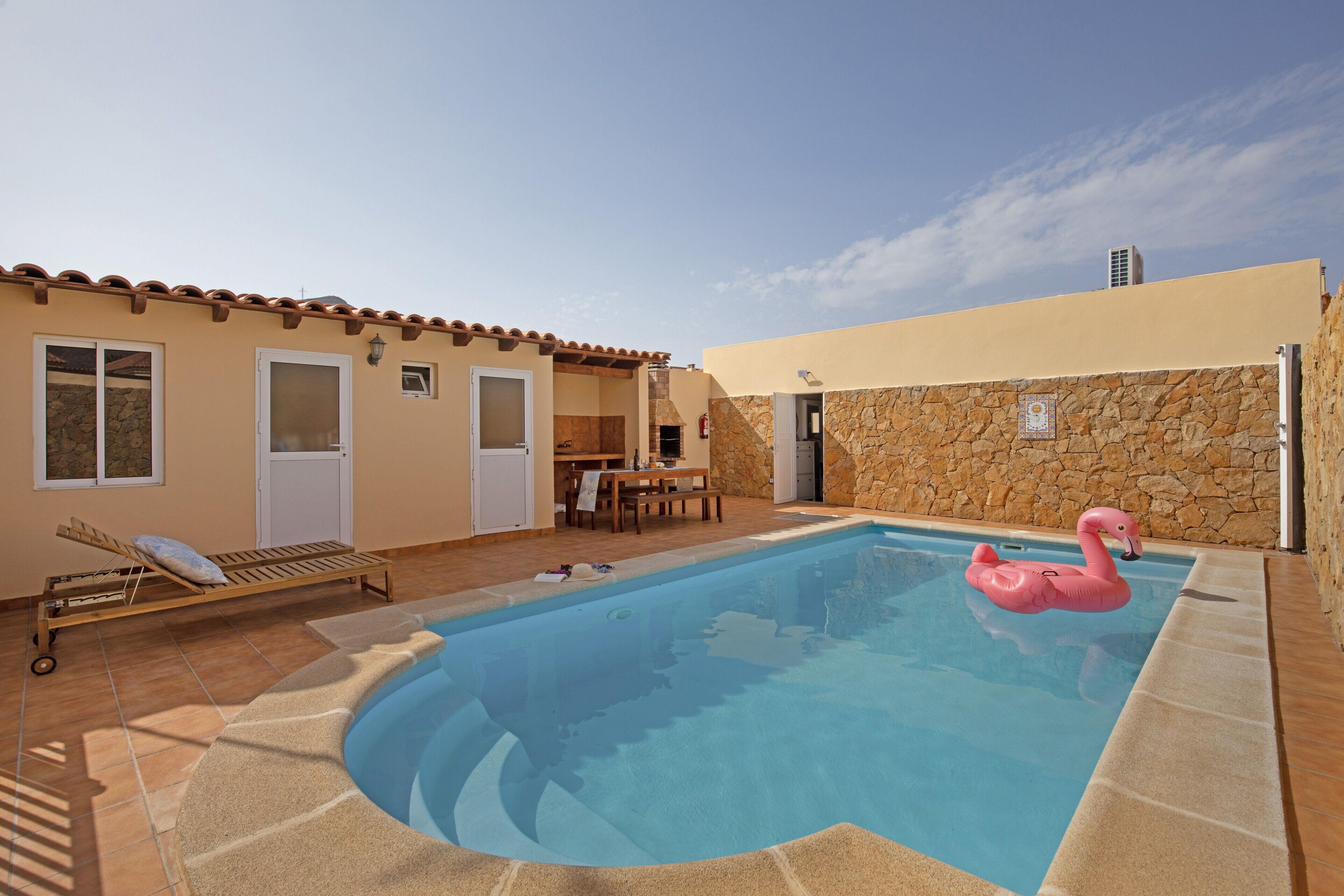 A swimming pool with a pink flamingo float, a sun lounger, and a stone wall.