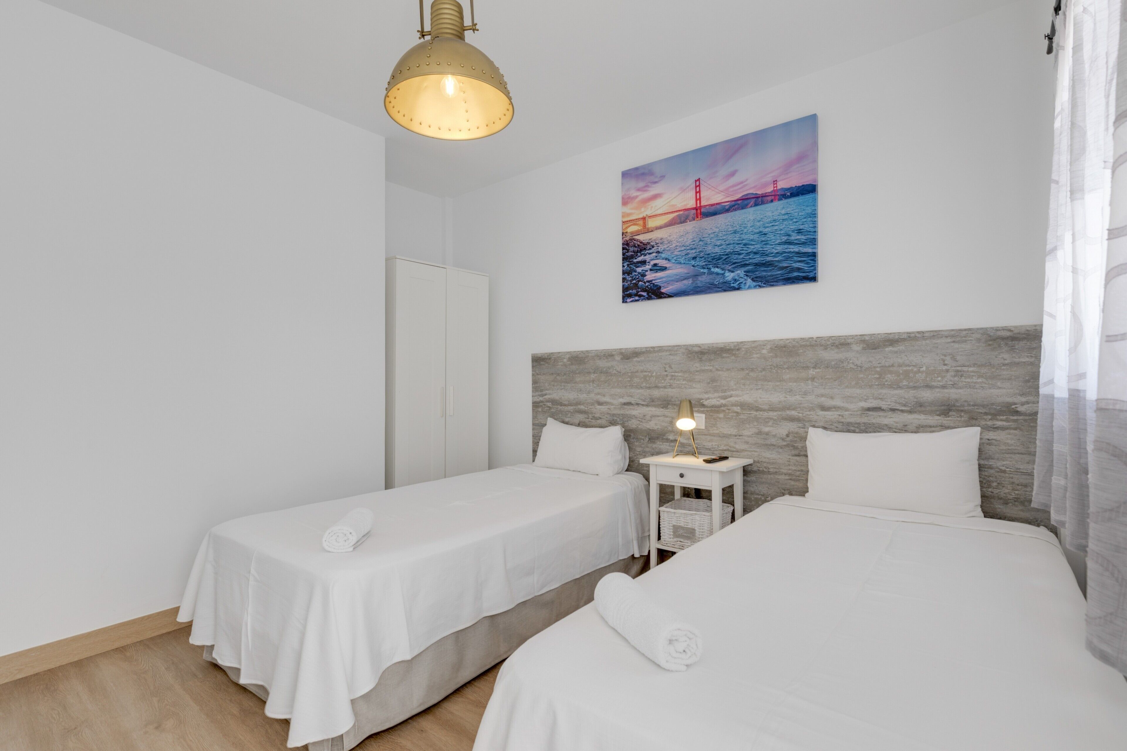 Una habitación de hotel con dos camas individuales, una mesita de noche con lámpara, un cuadro de un puente y un cabecero de madera.