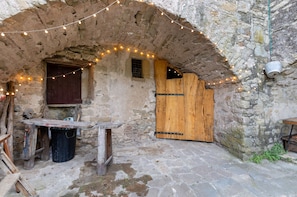 An arched stone building with a wooden door, a small window, and a table with a trash bin. - Chalet 'Casa Refugi Les Orenetes' with Shared Terrace and Kitchen (Rocafort)