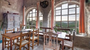 Un comedor rústico con una mesa de madera, sillas y un aparador. Se ven ventanas arqueadas grandes con cortinas.