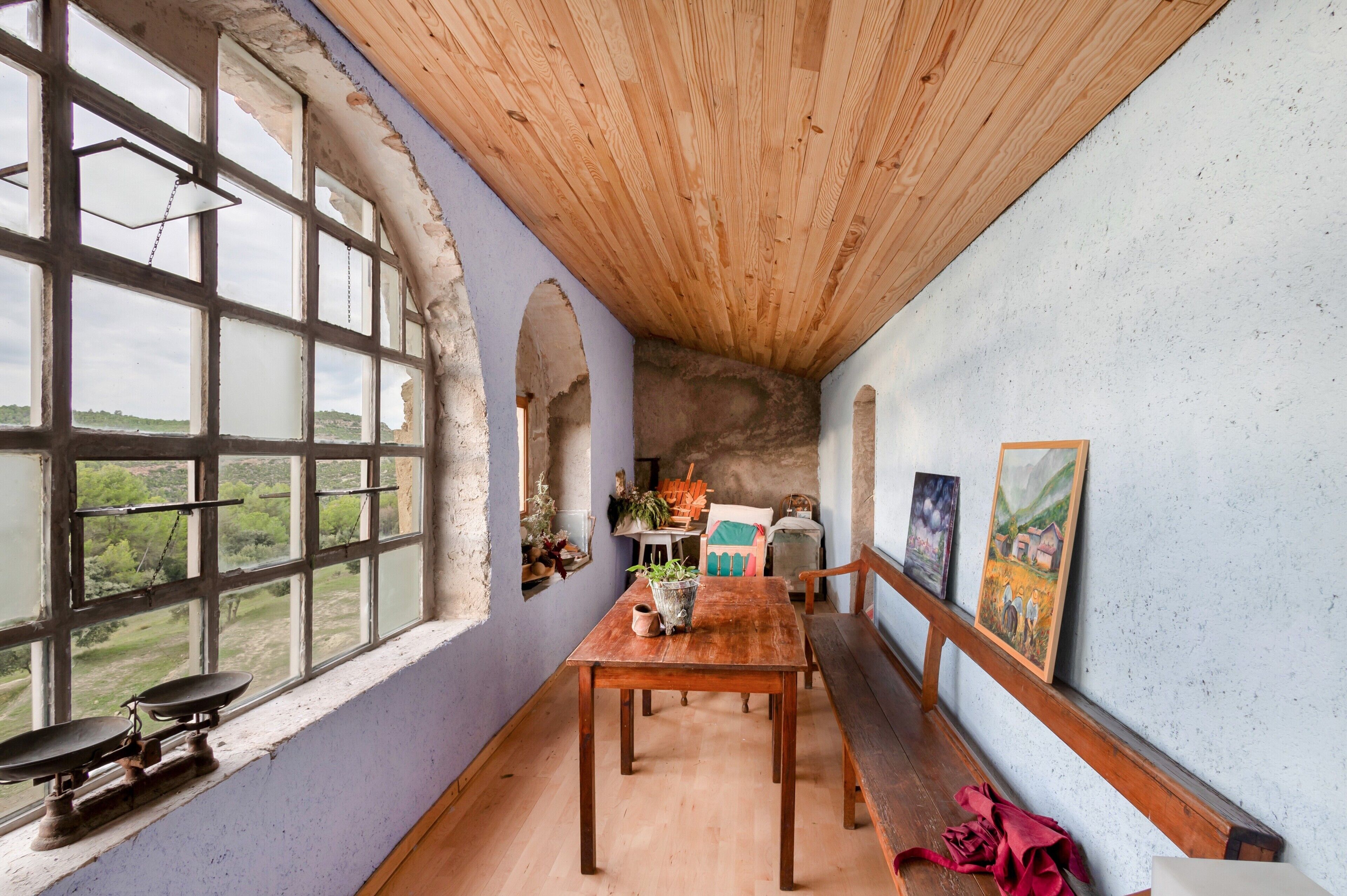 Ein rustikaler Raum mit Holzbalkendecke, einem großen Fenster mit Blick und einem Holztisch, sowie Bildern an den Wänden.