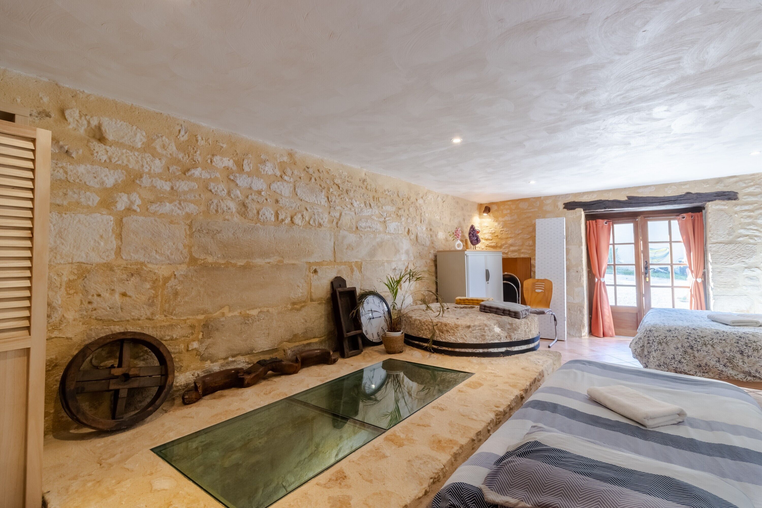 Una habitación con pared de piedra, una cama con ropa de cama a rayas, un armario de madera, un espejo redondo y una ventana con cortinas.