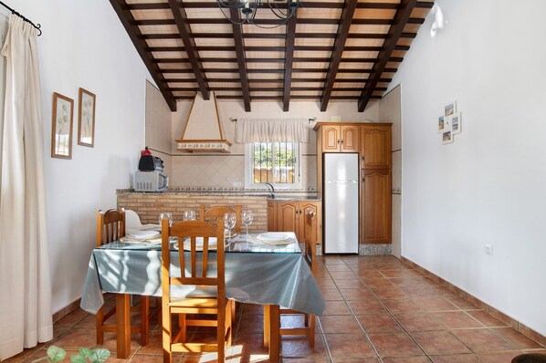 A dining area with a table set for two, wooden chairs, a kitchen with wooden cabinets, a refrigerator, and a window with a curtain.