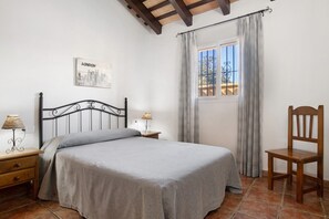 A bedroom with a bed, a wooden chair, a nightstand with a lamp, a window with curtains, and a wall-mounted picture of London.