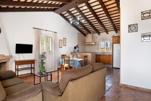 A living room with a sofa, TV, and fan, a dining area with a table and chairs, and a kitchen with cabinets and appliances.