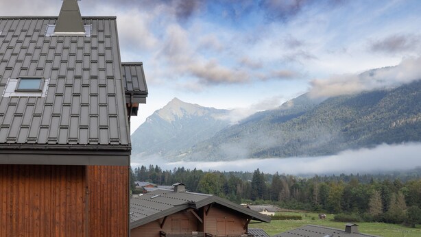 Une cabane de montagne avec un toit en pente, une façade en bois et une vue sur une vallée boisée, avec des montagnes en arrière-plan.