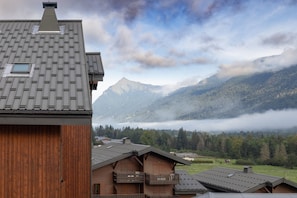 Una baita di montagna con tetto spiovente, facciata in legno e vista su una valle boscosa con montagne sullo sfondo.