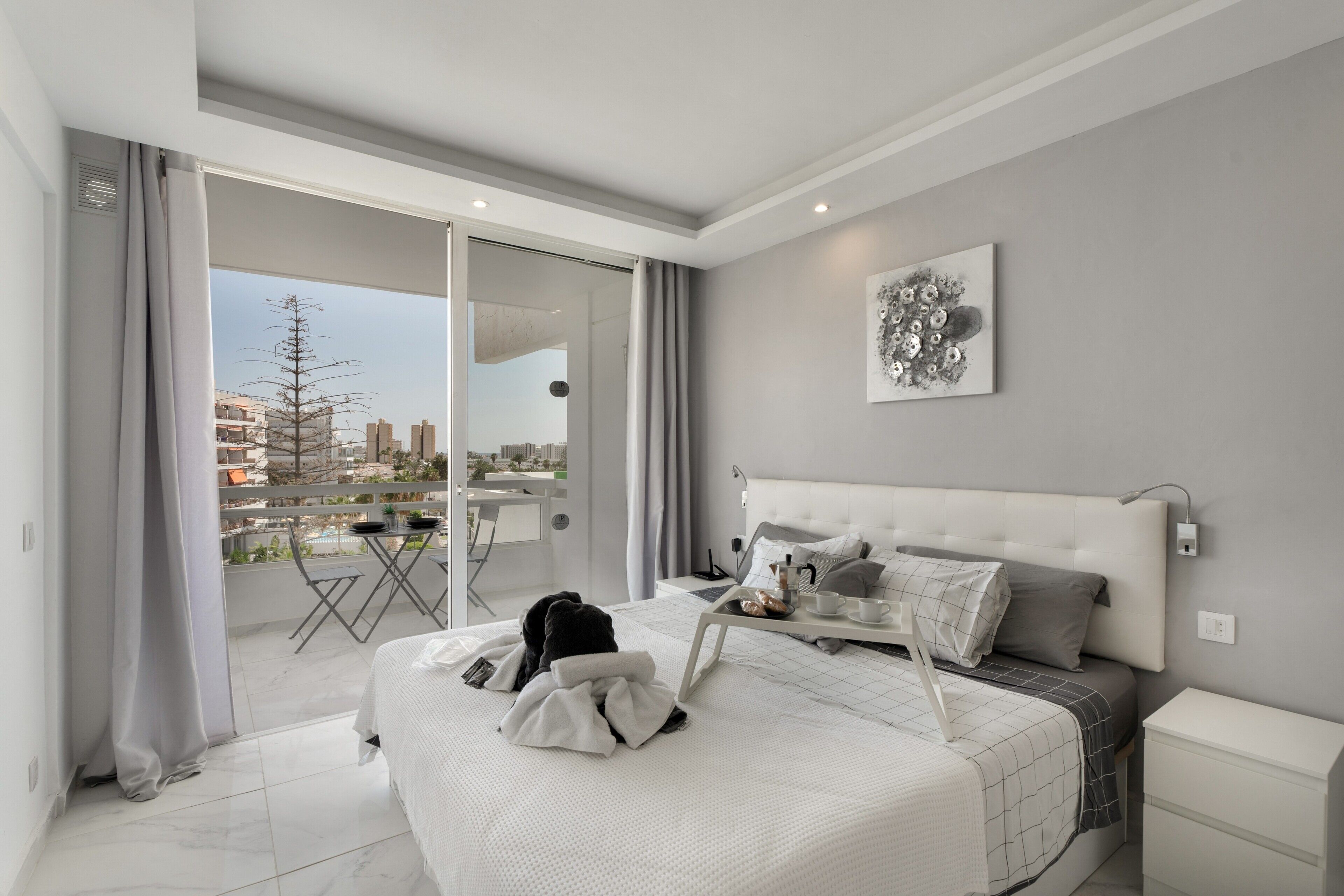 A modern bedroom with a large bed, a balcony with a view, and a cityscape visible through the glass door.