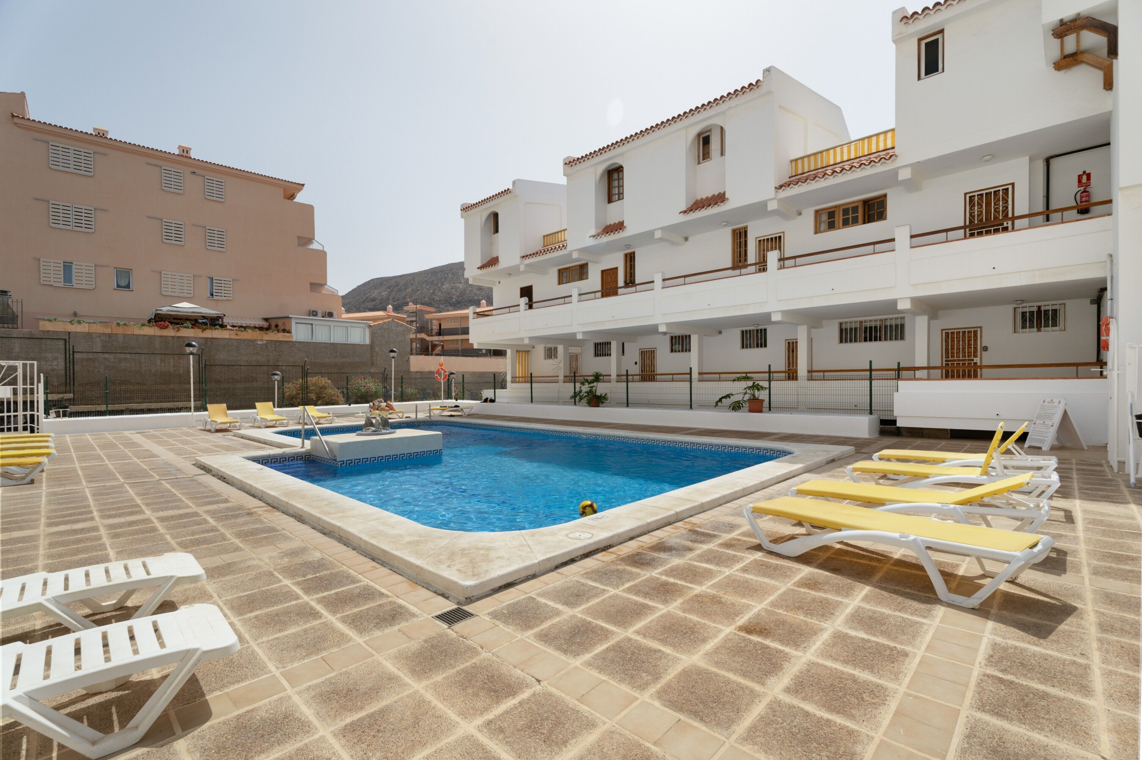 A swimming pool with sun loungers, a covered area, and a building with balconies.