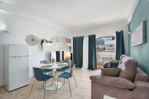 A modern kitchen with a refrigerator, dining table, and chairs. There is a sofa and a window with curtains.