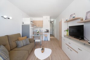 Un espace de vie compact comprenant un canapé, une petite table, un téléviseur et, au fond, une kitchenette.
