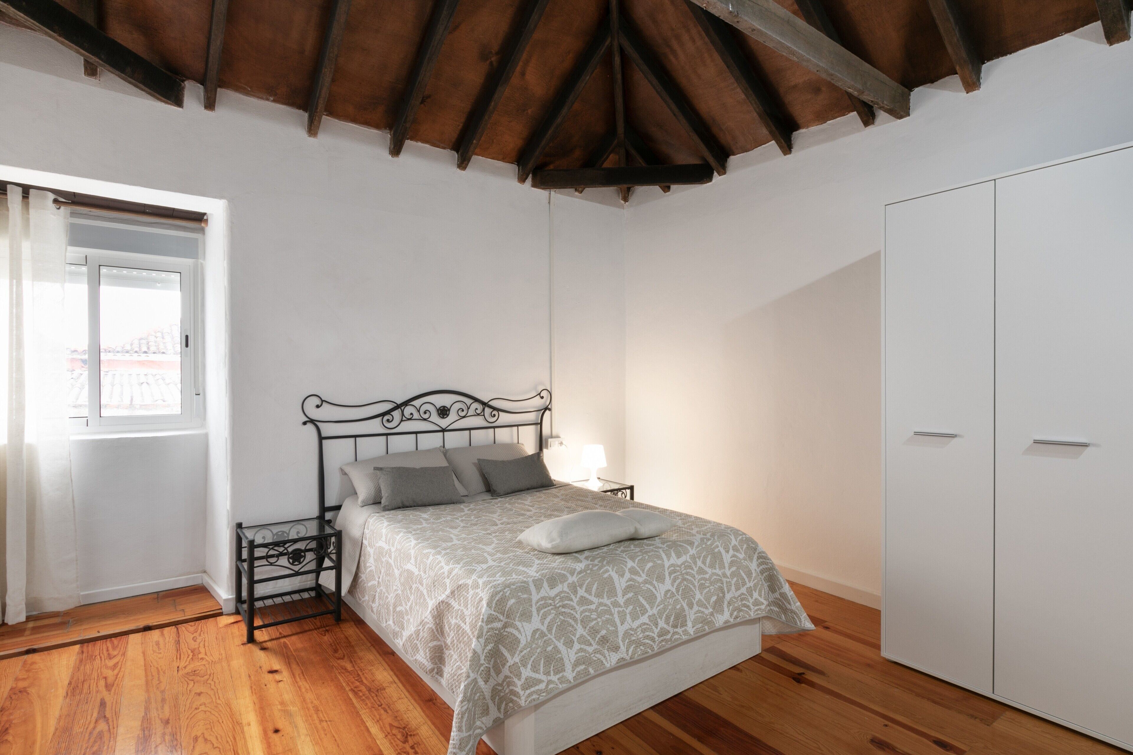 Een slaapkamer met een houten vloer, een bed met een beddeken met een patroon, een nachtkastje, een raam en een kledingkast.