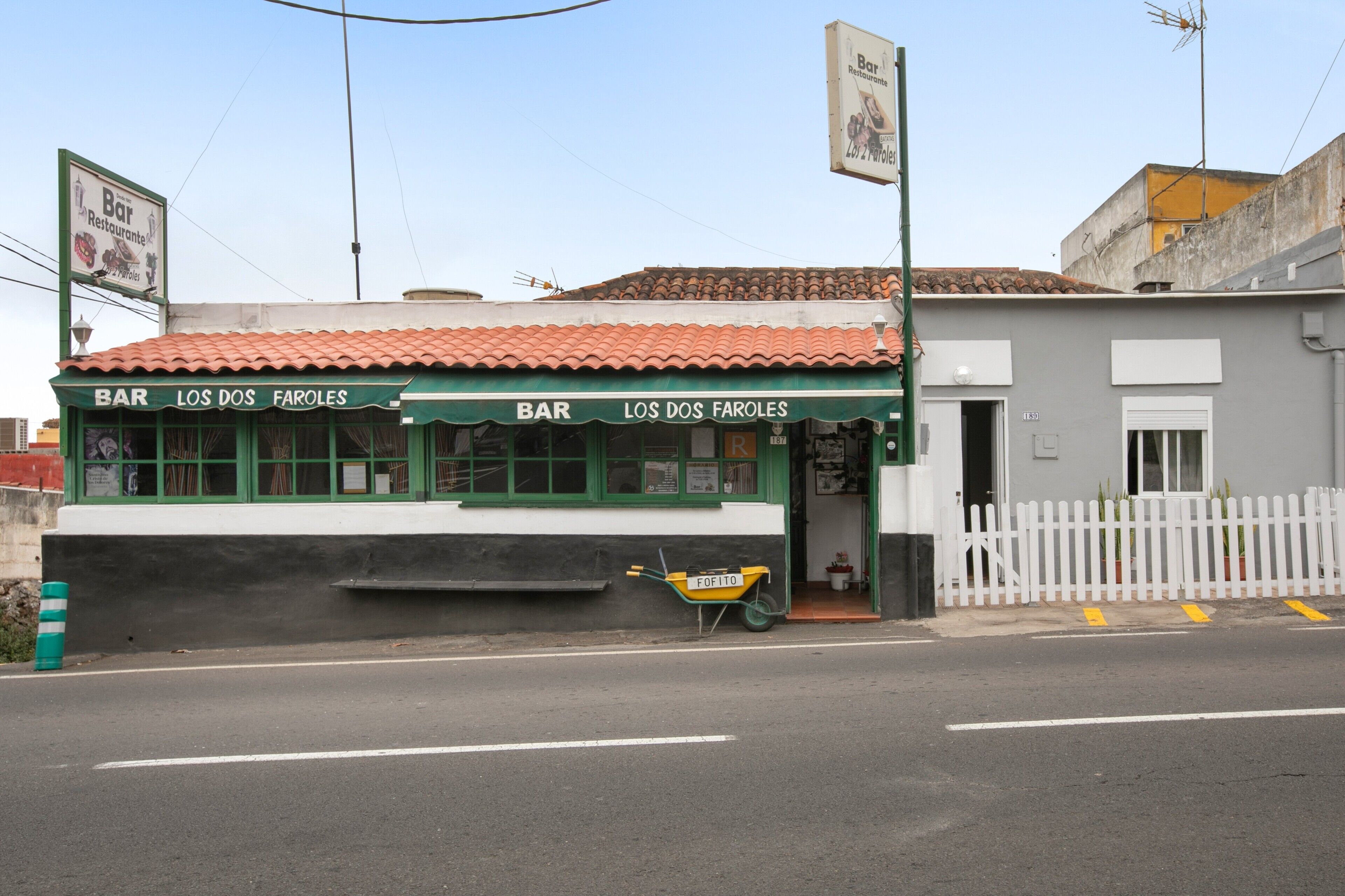 En bar som heter ”Los Dos Faroles” med en grön fasad, en skottkärra utanför och ett vitt trästaket.