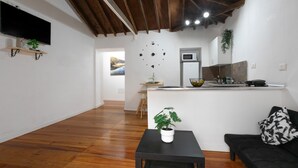 A modern living room with a wooden floor, a black sofa, a small table with a plant, a TV mounted on the wall, and a kitchen area in the background.