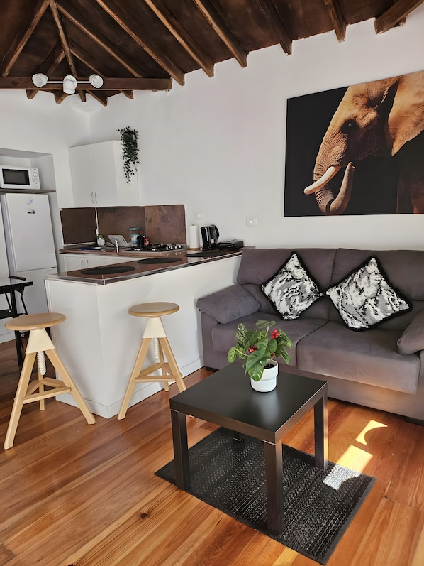A modern kitchen with a central island, wooden stools, a grey sofa with patterned cushions, and a large elephant artwork on the wall.