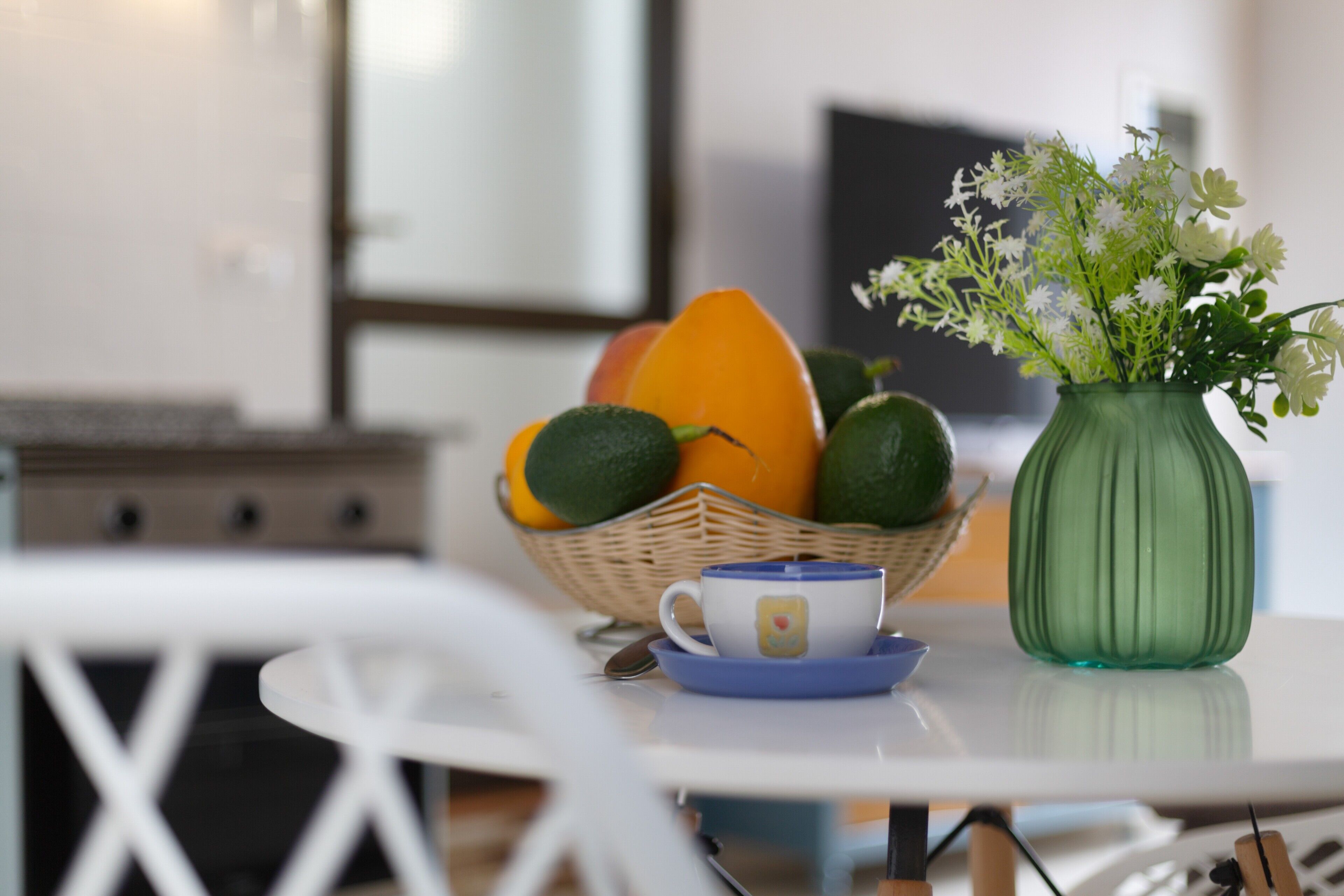 Una mesa de comedor con una cesta de frutas, una taza y un jarrón verde con flores.