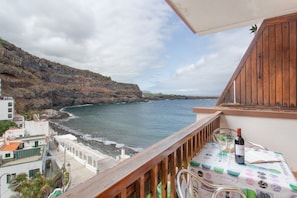 Een balkon met uitzicht op zee, een tafel met fles en glazen, en een gebouw met een schuine kap.