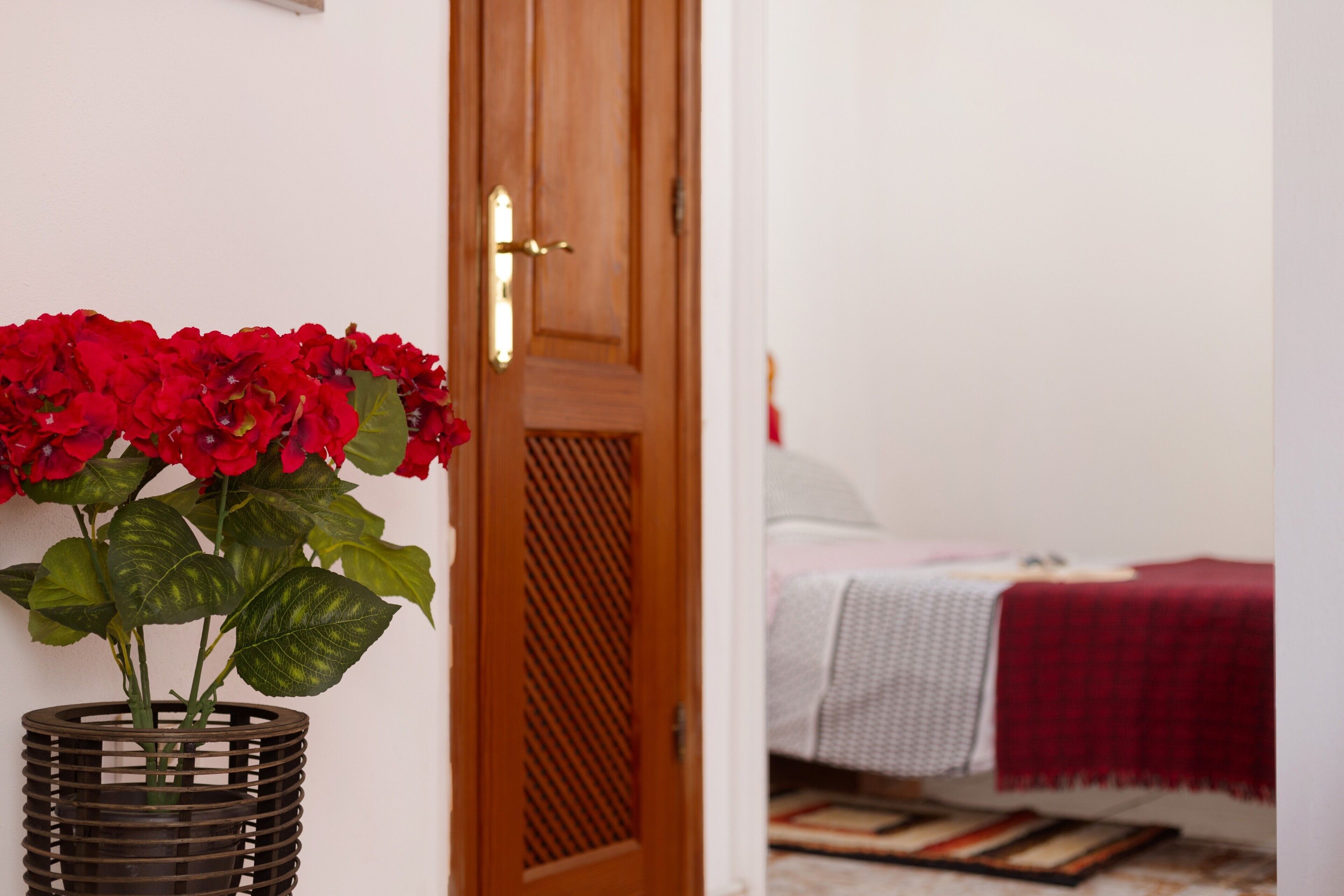 Una puerta de madera con manija de latón, una planta en maceta con flores rojas, y una cama con una colcha roja al fondo.