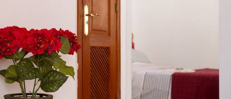 Una puerta de madera con manija de latón, una planta en maceta con flores rojas, y una cama con una colcha roja al fondo.