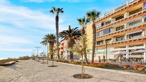 En anlagt promenade med palmer, en bygning med flere etager og altaner, samt udendørs siddepladser.